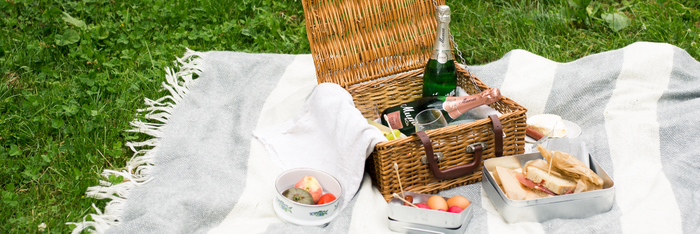 Auf einer gestreiften Decke liegen ein Picknickkorb, Gläser, frisches Obst und ein paar belegte Brote in einer Dose.