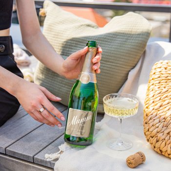 Eine Hand hält eine Flasche Mumm Sekt, während ein Glas mit sprudelndem Getränk auf einem Tisch steht.