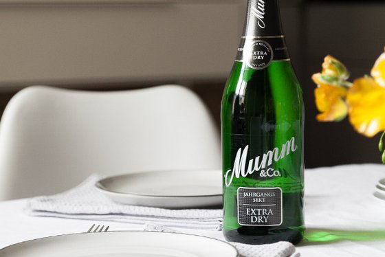 Eine Flasche Mumm Sekt steht auf einem gedeckten Tisch, umgeben von weißen Tellern und einer frischen Blumendekoration.