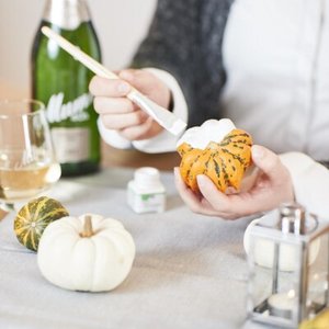 Eine Person dekoriert einen kleinen Kürbis mit einer weißen Farbe, während sie an einem festlich gedeckten Tisch sitzt.