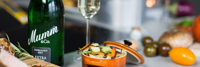 Eine Flasche Mumm Sekt steht neben einem Glas und einer bunten Obstschale auf einem Tisch, umgeben von frischen Zutaten.