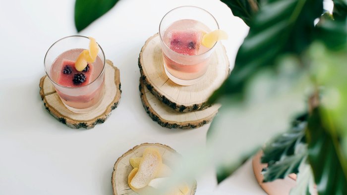 Die Gläser mit bunten Cocktails sind kunstvoll dekoriert und stehen auf rustikalen Holzuntersetzern, umgeben von grünen Blättern.
