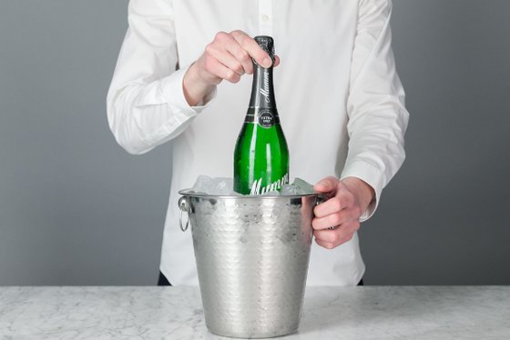 Eine Person hebt eine grüne Sektflasche aus einem Eimer voller Eis, der auf einem hellen Tisch steht.