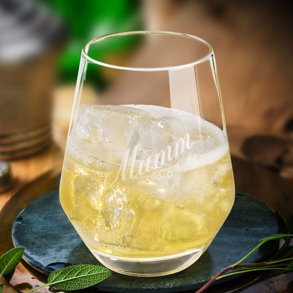 Das Glas mit dem Mumm-Logo enthält ein prickelndes Getränk, umgeben von frischen Kräutern und einer natürlichen Umgebung.