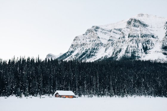 Eine kleine, rustikale Hütte steht in einer schneebedeckten Landschaft, umgeben von hohen, grünen Tannen und majestätischen Bergen.