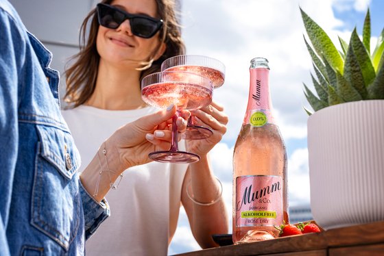 Eine Frau mit Sonnenbrille stößt mit einem Glas mit einer andere Person mit blauem Hemd an, daneben steht eine rosane Flasche Sekt und eine Pflanze