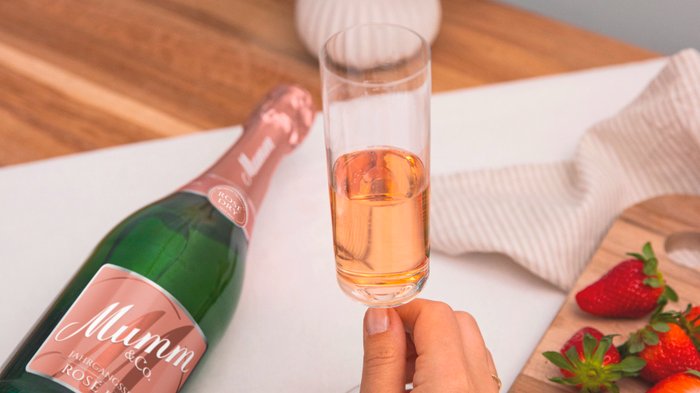 Ein Glas mit roséfarbenem Sekt wird von einer Hand gehalten, während eine Flasche Mumm daneben steht.