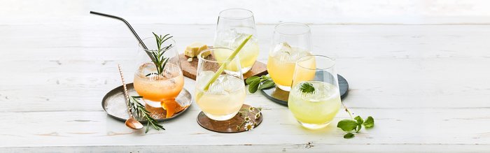 Bunte Cocktails in eleganten Gläsern, umgeben von frischen Kräutern und Zitrusfrüchten, auf einem hellen Holzuntergrund.