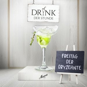 Auf einem weißen Tisch steht ein Glas mit einem leuchtend grünen Drink, umgeben von Eiswürfeln und einem kleinen Schild.