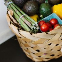 Ein geflochtener Korb voller frischer Gemüse und Obst, darunter Avocados, Paprika und Kirschtomaten, auf einem dunklen Tisch.