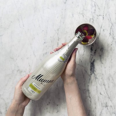 Die Hand einer Person hält eine Flasche Mumm alkoholfrei, während eine Schale mit Erdbeeren und Himbeeren daneben liegt.
