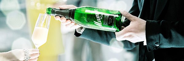 Ein stilvoll gekleideter Mann reicht einer Person ein Glas Sekt, während er aus einer Flasche einschenkt, umgeben von festlicher Atmosphäre.