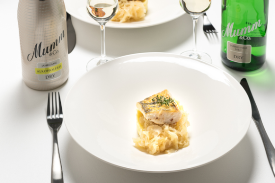 Auf dem Tisch steht ein kunstvoll angerichteter Teller mit Fisch und Sauerkraut, begleitet von zwei Sektgläsern und Flaschen.