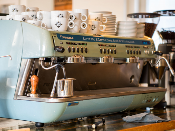 Eine elegante, mintgrüne Espressomaschine steht auf einer Holztheke, umgeben von weißen Kaffeetassen und Kaffeebohnen.