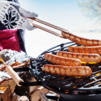 Eine Person mit einem Handschuh grillt Würstchen auf einem Grill, während der Schnee im Hintergrund liegt.