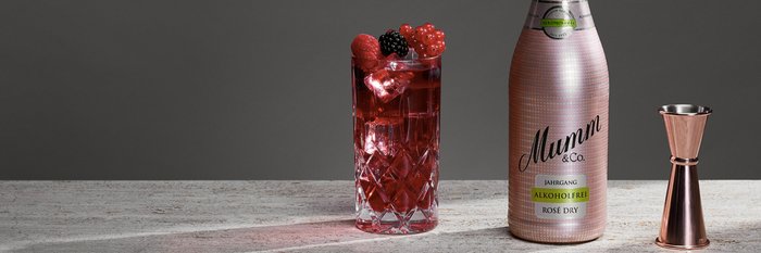 Eine Flasche alkoholfreier Rosé-Sekt neben einem dekorativen Glas voller Eis und frischen Beeren, auf einem hellen Tisch platziert.