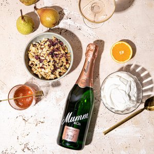 Ein eleganter Tisch mit einer Flasche Mumm Rosé, frischen Birnen, Joghurt und einer Schüssel mit Müsli, umgeben von frischen Früchten.