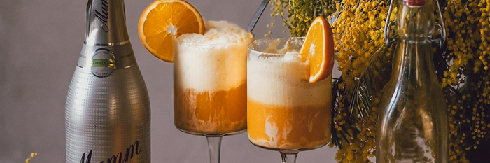 Zwei Gläser mit schaumigem, orangefarbenem Getränk, garniert mit einer Orangenscheibe, stehen neben einer Flasche Sekt.