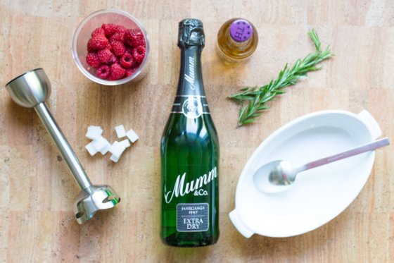 Die Szene zeigt eine grüne Sektflasche, frische Himbeeren in einer Schüssel und Zutaten für einen Cocktail auf einem hellen Tisch.