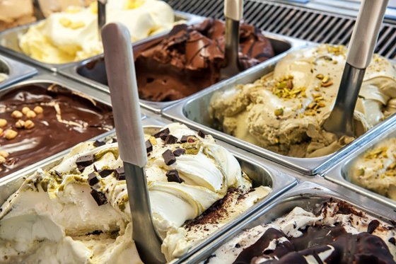Eine Auswahl an verschiedenen Eissorten in einer Eisdiele, darunter Schokolade, Pistazie und Vanille, mit Löffeln zum Servieren.