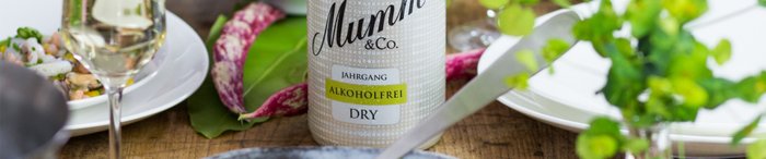 Auf einem rustikalen Holztisch stehen ein Glas Sekt, eine Flasche alkoholfreier Sekt und ein Teller mit feinem Essen.