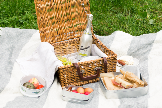 Ein geflochtener Picknickkorb steht auf einer Decke, gefüllt mit Gläsern, einer Flasche alkoholfreiem Sekt und frischen Snacks.