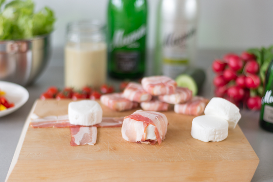 Eine appetitliche Anordnung von Speck umwickelten Käseportionen auf einem Schneidebrett, mit frischem Gemüse im Hintergrund.
