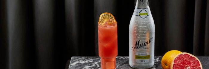 Eine Flasche Mumm alkoholfrei steht neben einem bunten Cocktail und frischen Zitrusfrüchten auf einem marmorierten Tisch.