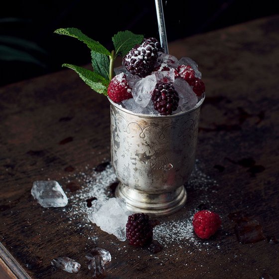 Ein silberner Becher gefüllt mit zerstoßenem Eis, frischen Himbeeren und Brombeeren, umgeben von Eisstücken auf einem Holztisch.