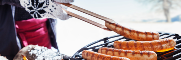 Eine Person mit einem gestrickten Handschuh greift mit einer Zange nach gegrillten Würstchen auf einem Grill im Freien.