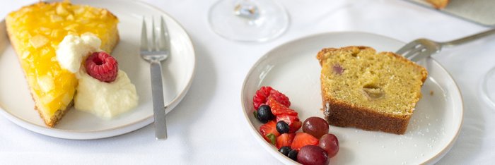 Ein Stück Ananaskuchen mit Sahne und Himbeere, daneben ein Stück saftiger Kuchen mit frischen Beeren auf einem weißen Tisch.