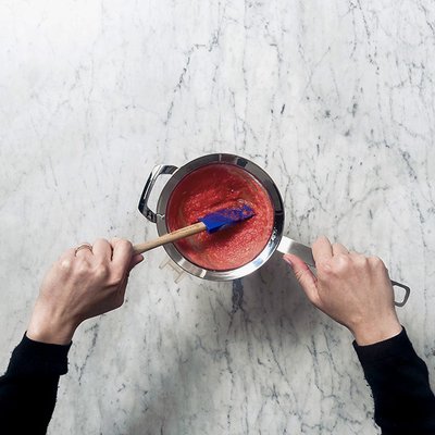 Eine Person rührt mit einem blauen Spatel in einem silbernen Topf mit roter Soße auf einer marmorierten Arbeitsplatte.