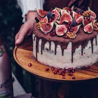 Eine üppige Torte mit Schokoladenglasur, dekoriert mit frischen Feigen und Granatapfelkernen, wird auf einem Holzbrett präsentiert.